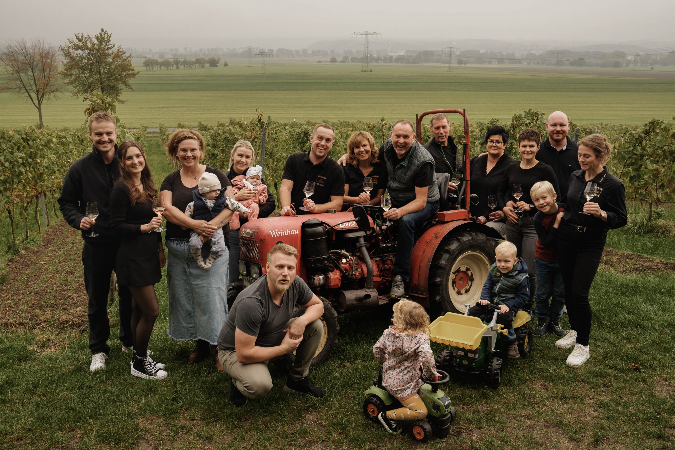 Eine große Gruppe von Menschen, darunter Erwachsene und Kinder, versammelt sich fröhlich um einen roten Traktor auf dem Weingut Steffen Loose in einem grasbewachsenen Weinberg. Einige halten Getränke in der Hand, Kleinkinder spielen auf Spielzeugfahrzeugen, und grüne Felder erstrecken sich in der nebligen Ferne hinter ihnen.
