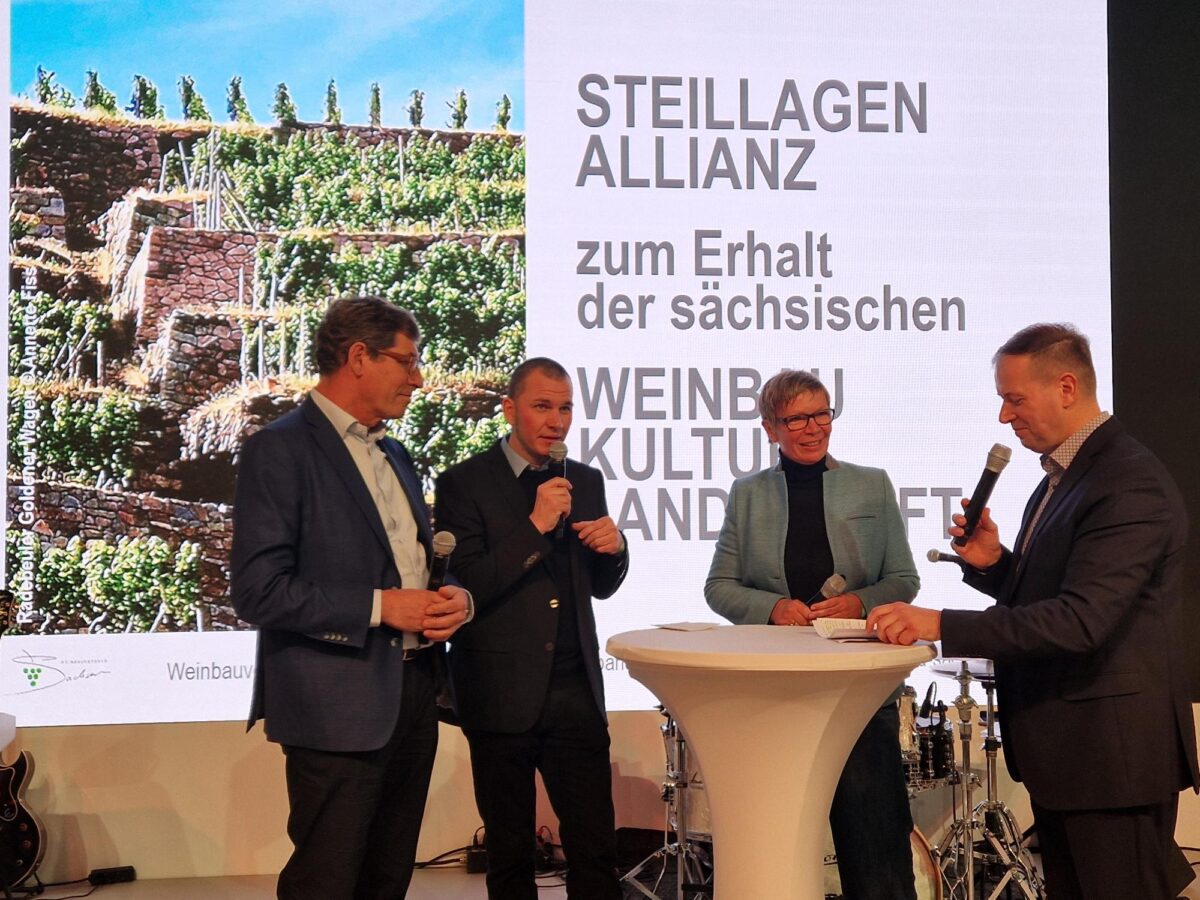 Vier Personen sprechen auf der Bühne bei einer Podiumsdiskussion mit Mikrofonen. Hinter ihnen zeigt eine Leinwand Weinbergterrassen und einen deutschen Text über die Erhaltung der sächsischen Weinkultur, in dem die Bedeutung der Steillagen und das fortwährende Engagement der Steillagenallianz hervorgehoben werden. Im Hintergrund ist Musikequipment zu sehen.